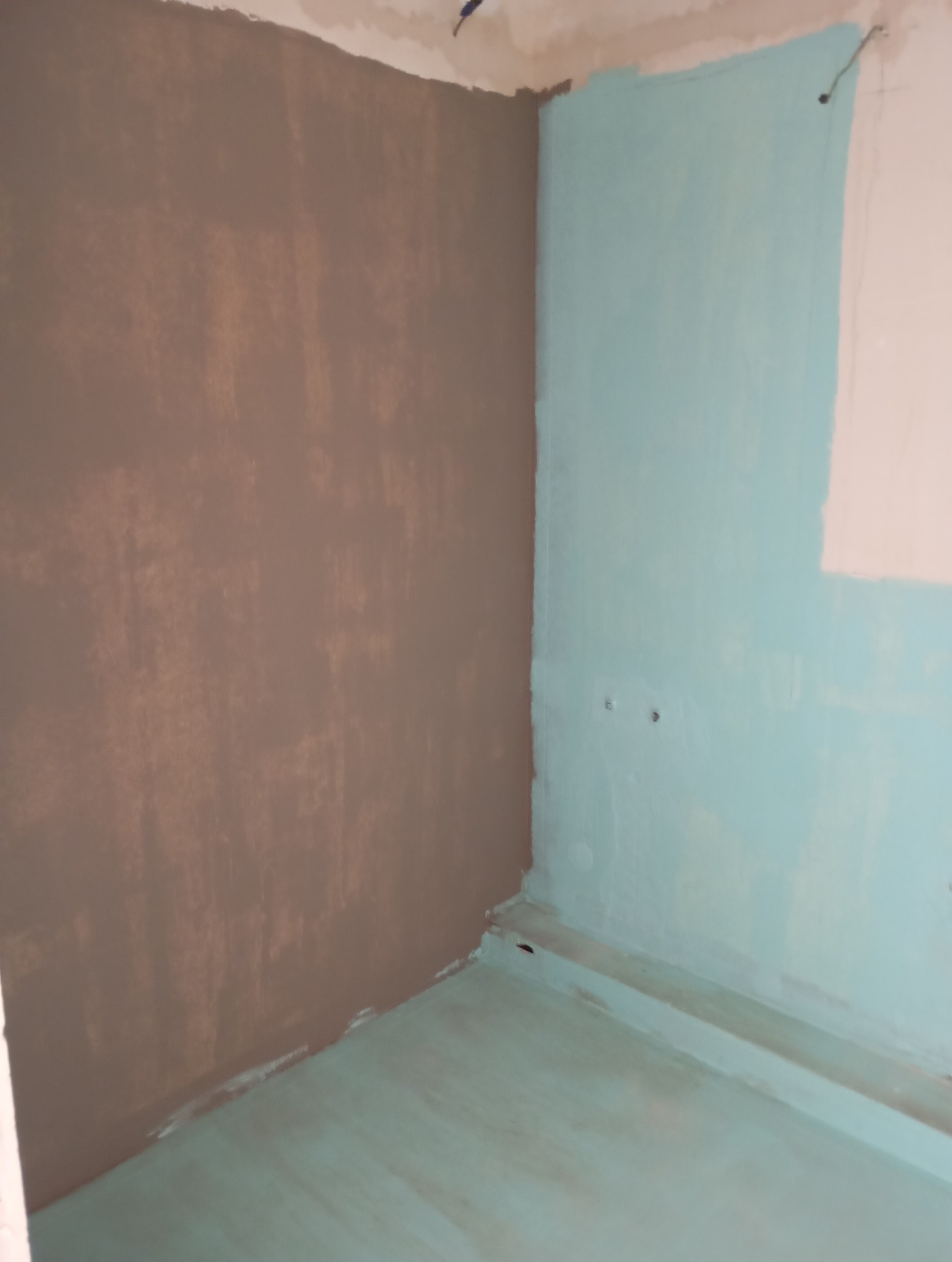 Narożnik pokoju w trakcie malowania: jedna ściana pokryta brązową farbą, druga jasnoniebieską, podłoga również pomalowana na jasnoniebiesko, widoczne niedociągnięcia malarskie.
