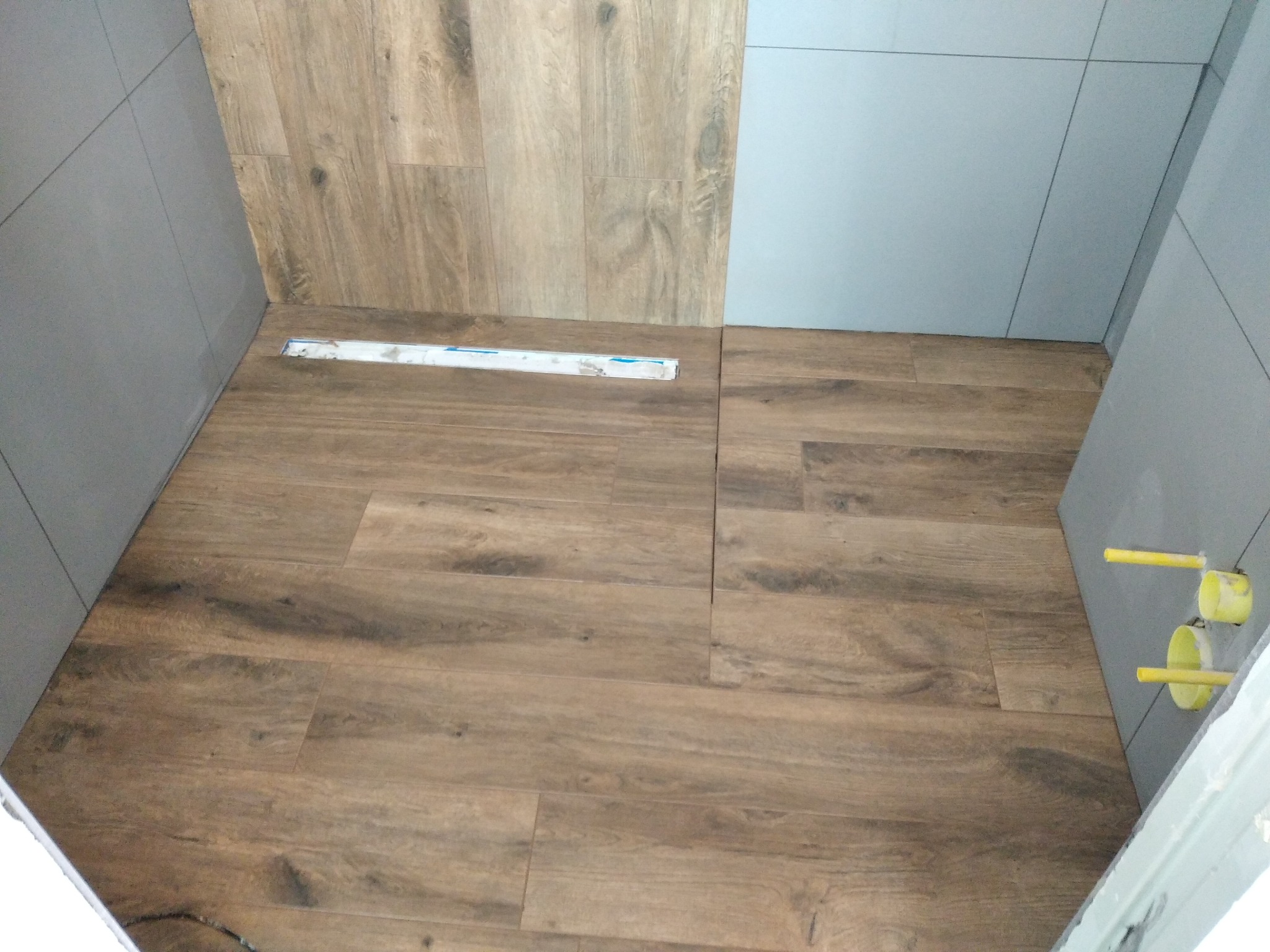 Ułożona podłoga z paneli imitujących drewno w łazience, widoczny odpływ liniowy i fragment ściany wyłożonej szarymi płytkami, żółte rury wystające ze ściany.