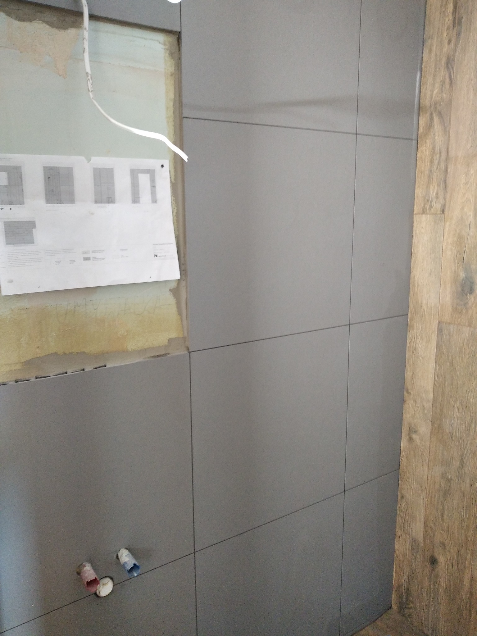 Ściana łazienki w trakcie prac: szare płytki ceramiczne ułożone do pewnej wysokości, odsłonięty fragment ściany z instalacją elektryczną i przyklejonym planem technicznym, fragment drewnopodobnej...