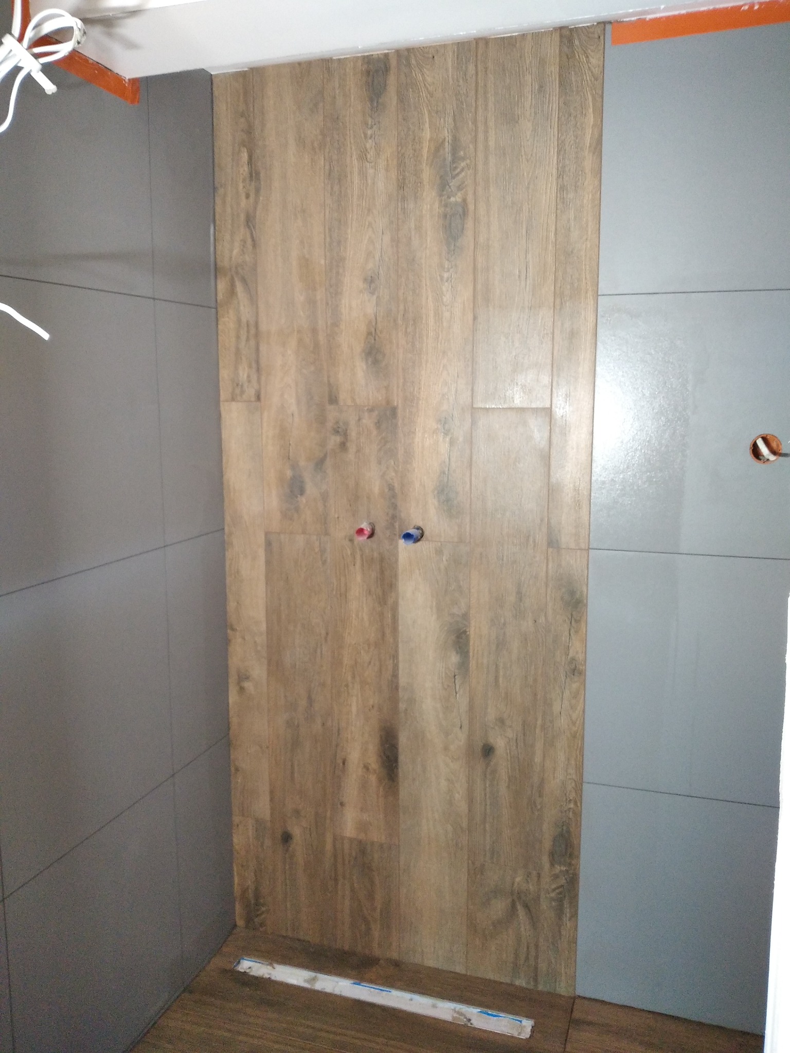 Łazienka w trakcie wykańczania: szare płytki ścienne zestawione z panelami imitującymi drewno, widoczne wystające rury i poziomica na podłodze.