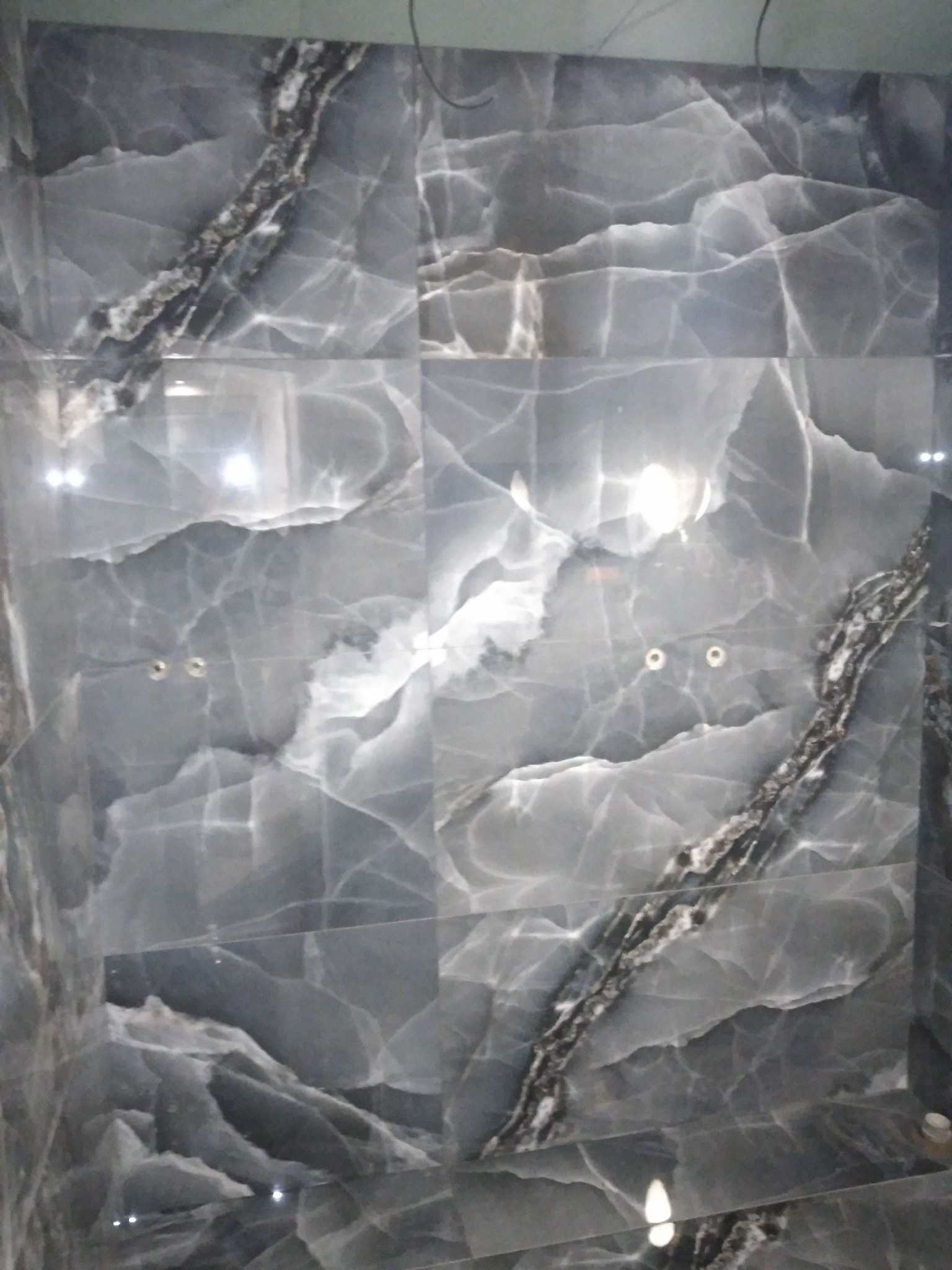 Ściana wyłożona szarymi płytkami imitującymi marmur z ciemnymi żyłkami, widoczne otwory na baterię.