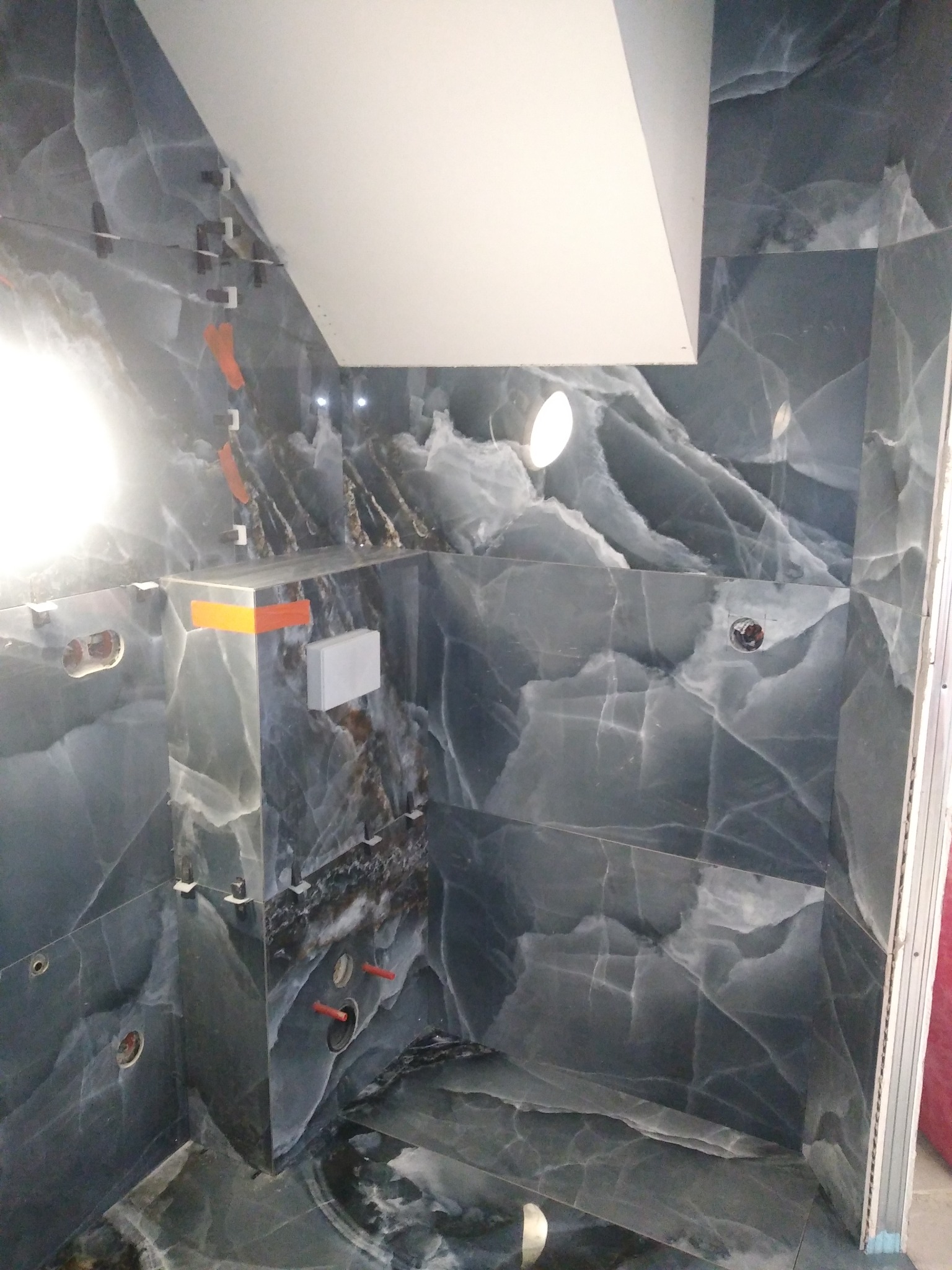 Montaż płytek imitujących marmur w kabinie prysznicowej, widoczne poziomowanie z użyciem klipsów i klinów, przygotowane otwory na armaturę, sufit podwieszany z oświetleniem.