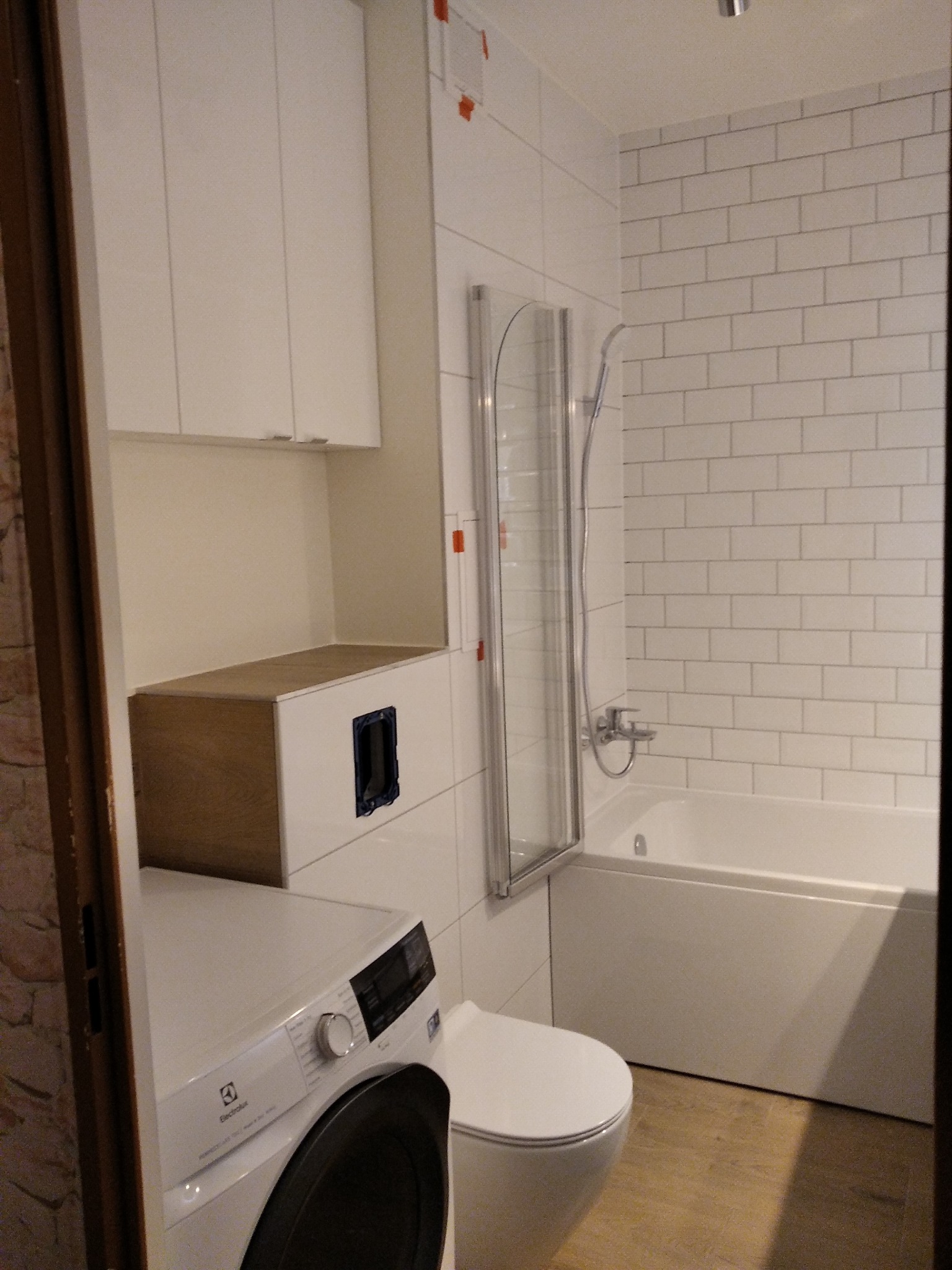 Nowoczesna łazienka z białą armaturą, kabiną prysznicową ze srebrnymi profilami, pralką Electrolux i białymi szafkami wiszącymi.