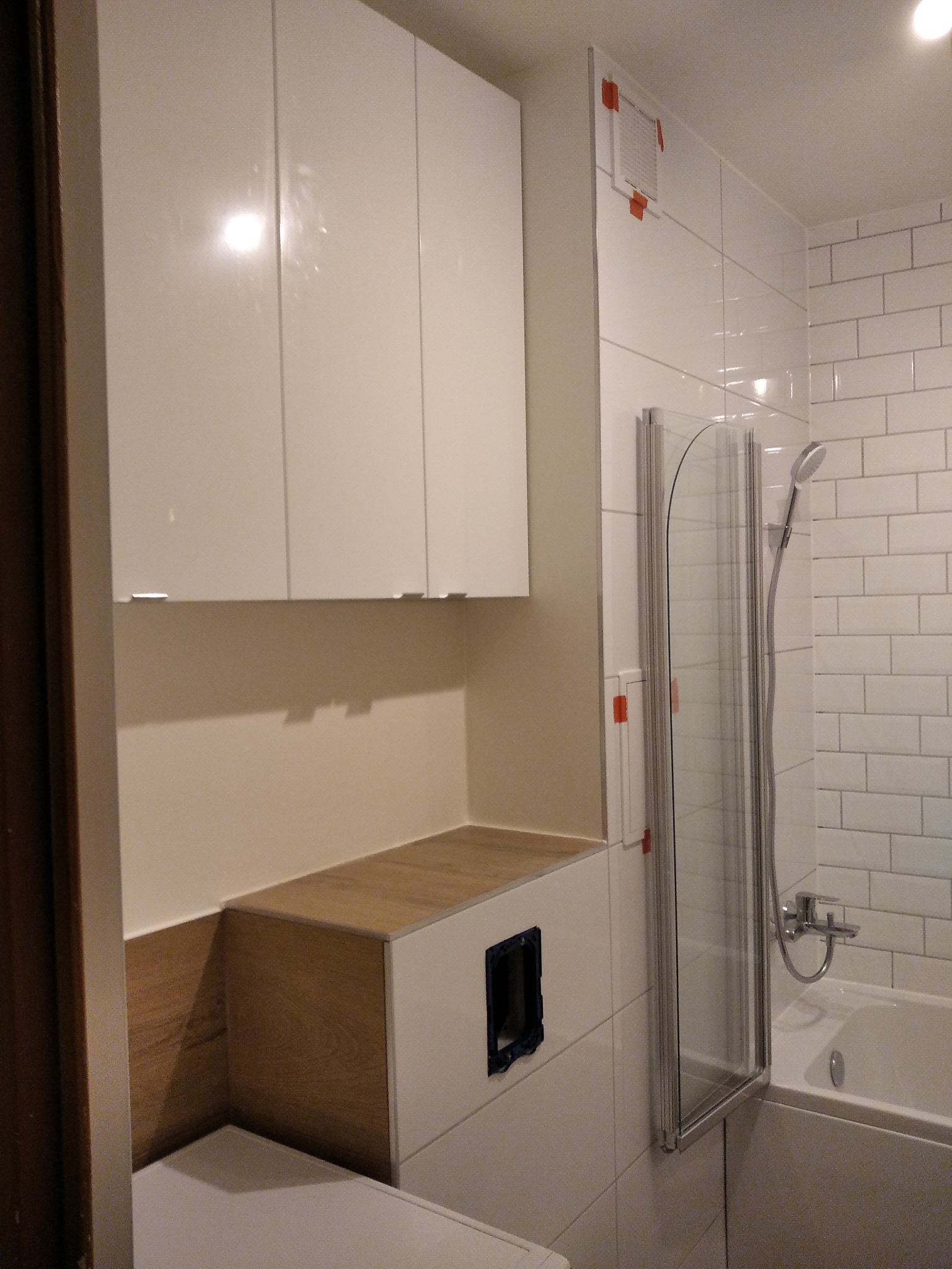 Biała łazienka z szafką wiszącą, zabudową WC z drewnopodobnym blatem i kabiną prysznicową z pojedynczymi drzwiami, widoczna instalacja elektryczna przed montażem osprzętu.