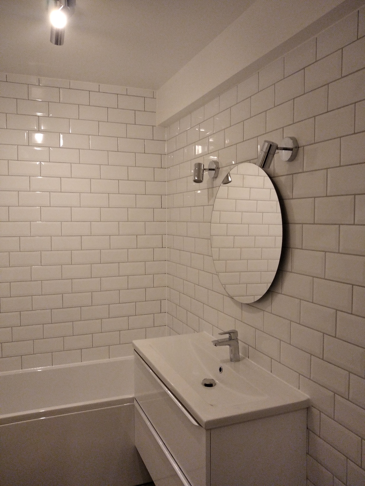 Biała łazienka z wanną, umywalką z szafką i okrągłym lustrem, wykończona białymi, prostokątnymi płytkami ułożonymi w cegiełkę.