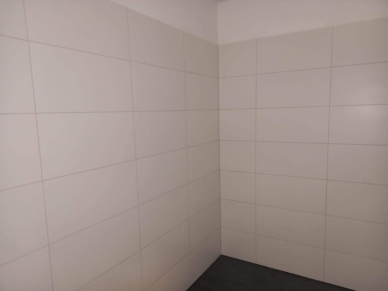 Ściana wyłożona białymi, matowymi płytkami w układzie prostokątnym z ciemną fugą i fragmentem szarej podłogi.