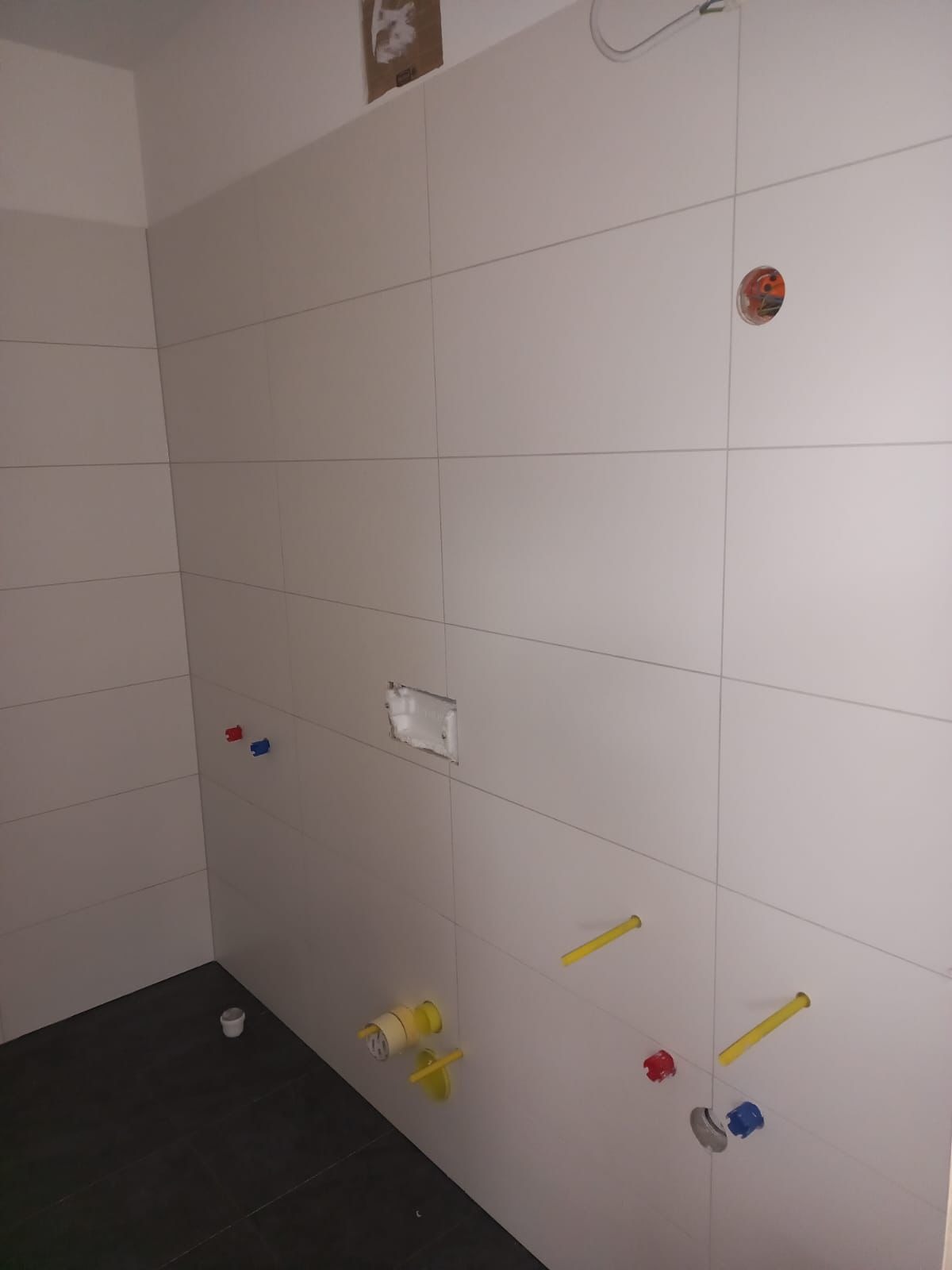 Świeżo wykafelkowana ściana łazienki z wystającymi rurami instalacyjnymi i puszką elektryczną w przygotowaniu do montażu armatury.