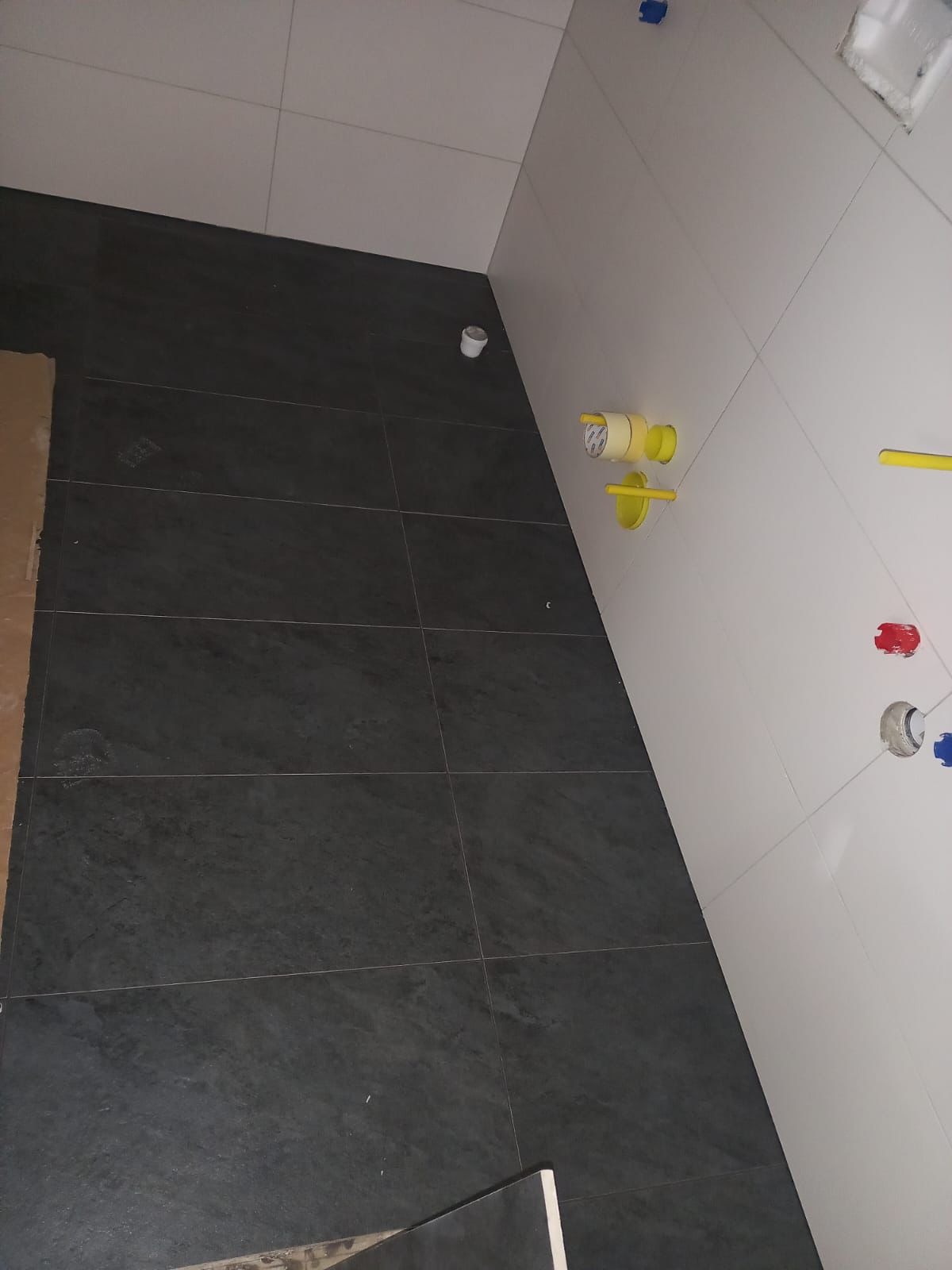 Ciemnoszara, matowa podłoga z płytek w trakcie prac wykończeniowych, widoczne przygotowane przyłącza wodne na białej ścianie i fragment kartonu z płytkami.
