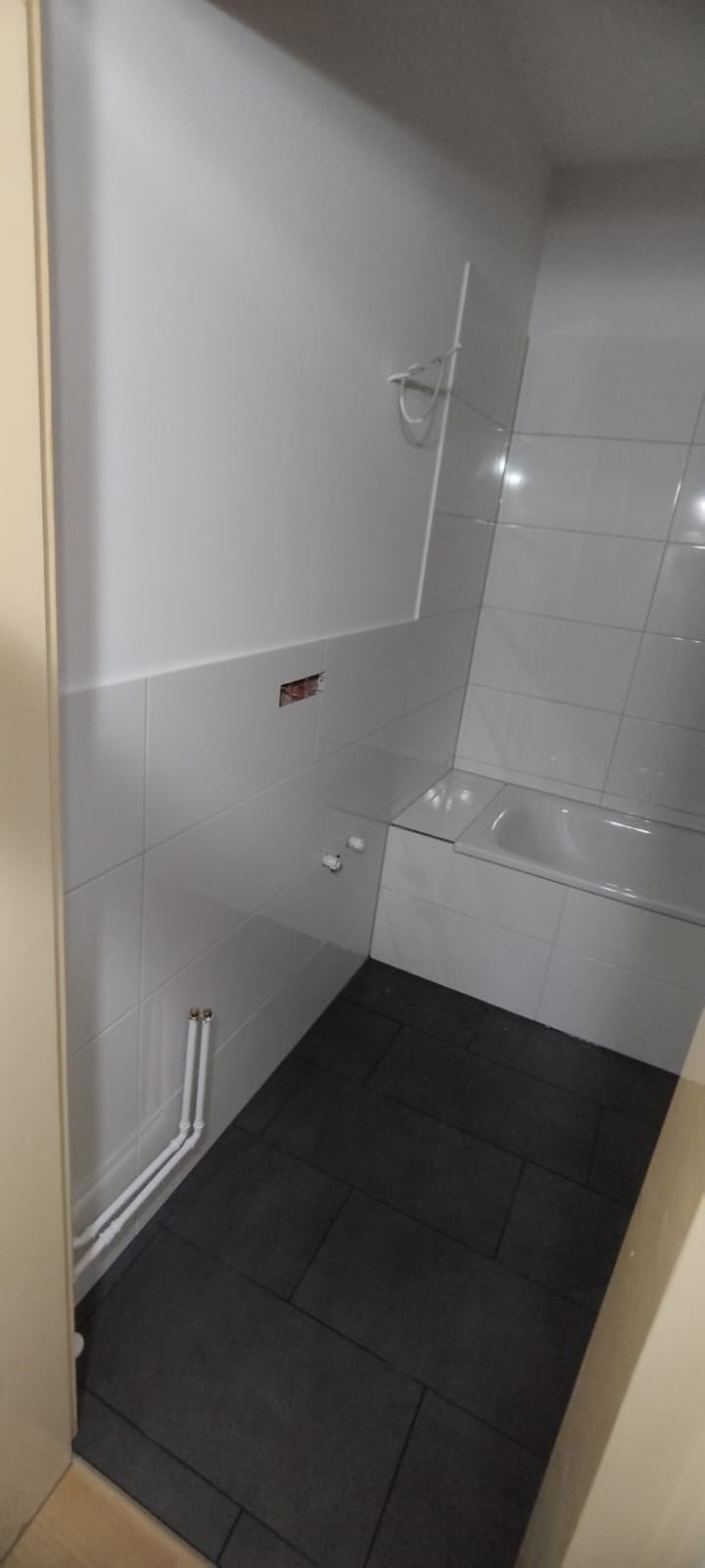 Białe płytki ścienne w łazience, częściowo ułożone, z widocznymi rurami i otworem na gniazdko elektryczne, oraz ciemne, grafitowe płytki podłogowe ułożone w jodełkę, fragment wanny w rogu.