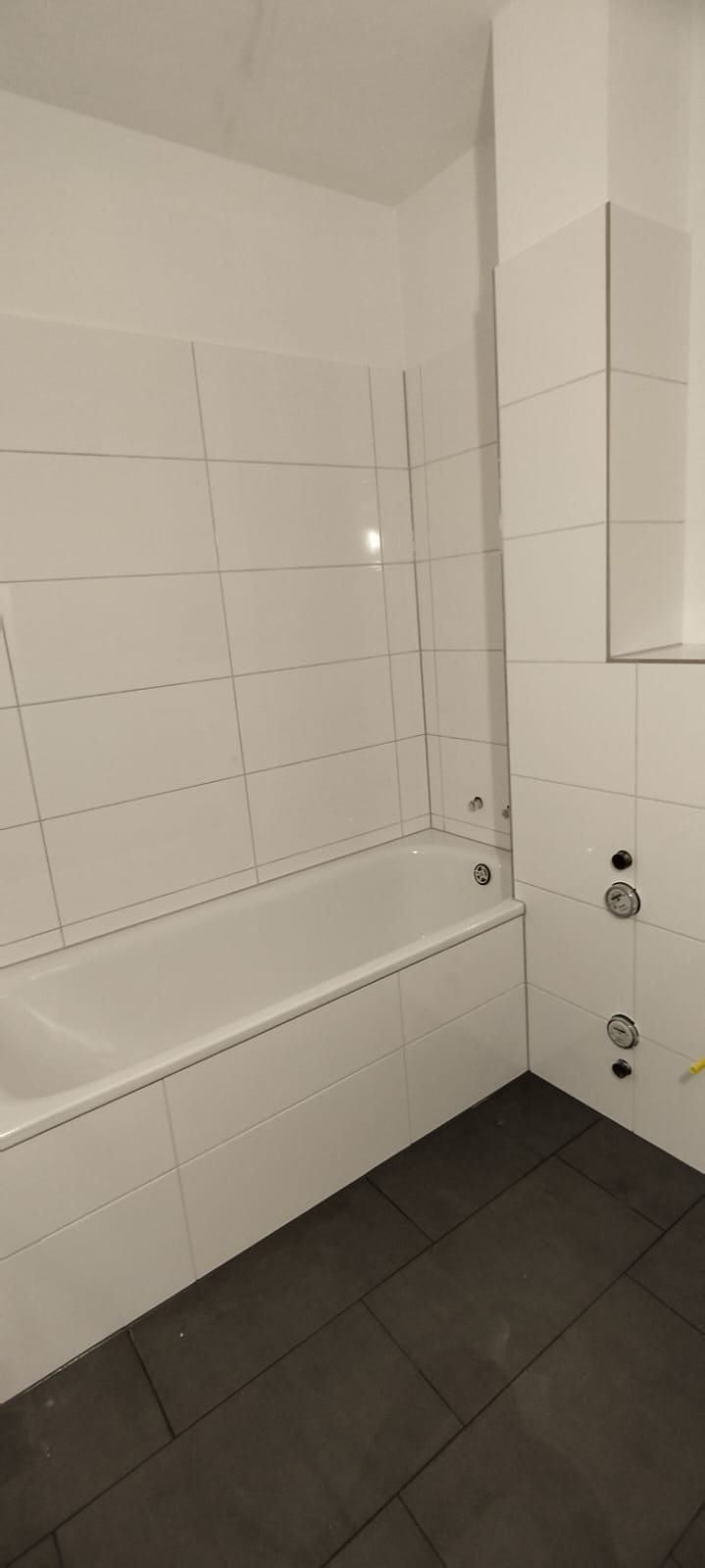 Biała wanna obudowana płytkami, z widocznymi zaworami i ciemną podłogą w łazience po remoncie.