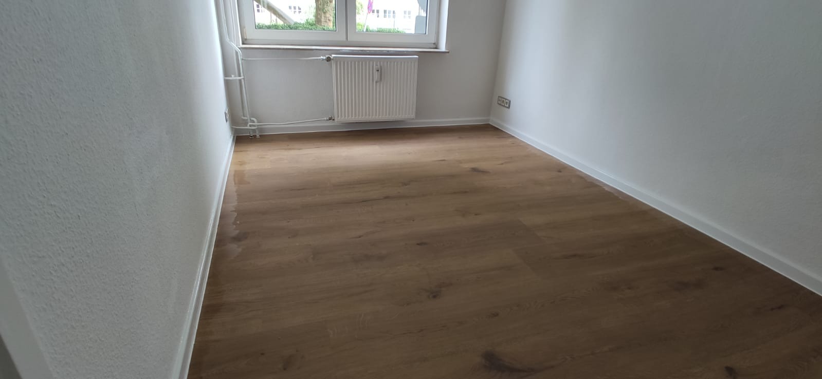 Pusty pokój z nową podłogą z paneli w odcieniach brązu, białe ściany, grzejnik pod oknem. Widoczne listwy przypodłogowe.