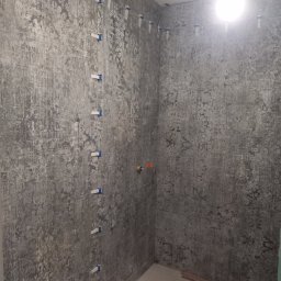 Apres-Bud Hennadii Zalizovskyi - Ściany łazienki w trakcie układania płytek o wzorze imitującym beton, z użyciem systemu poziomowania płytek z niebieskimi klipsami, widoczny fragment instalacji wodnej.