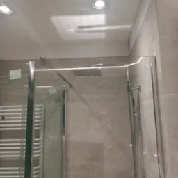 Nowoczesna kabina prysznicowa typu walk-in z chromowanymi profilami i deszczownicą, widoczny grzejnik drabinkowy w tle, beżowe płytki imitujące marmur na ścianach.