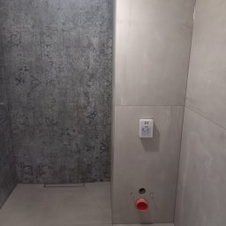 Apres-Bud Hennadii Zalizovskyi - Wnętrze łazienki w trakcie remontu, widoczne szare płytki na ścianach, podtynkowa instalacja wodno-kanalizacyjna z czerwoną rurą, oraz odpływ liniowy w podłodze.