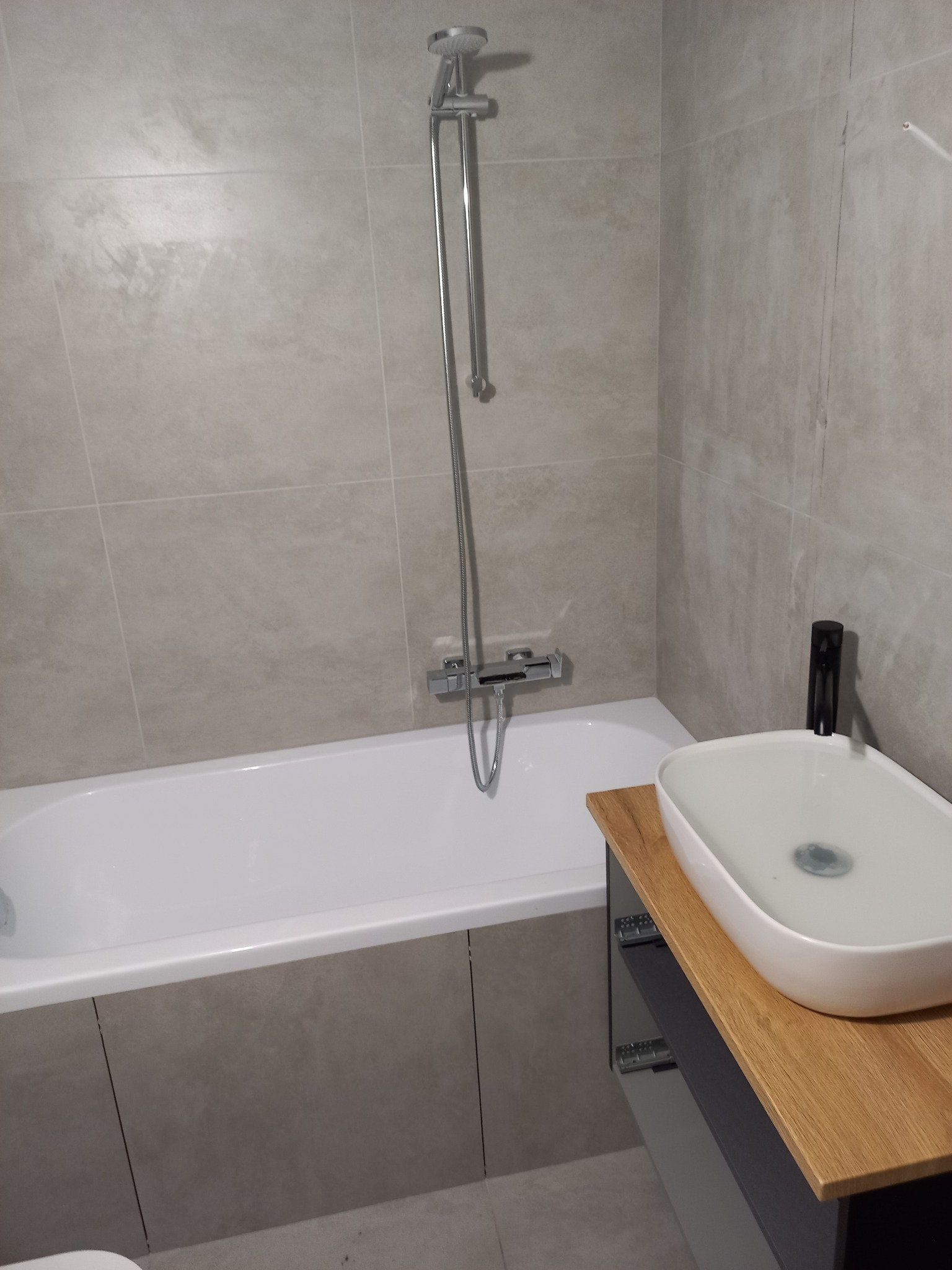 Nowoczesna łazienka z beżowymi płytkami, białą wanną obudowaną płytkami, umywalką nablatową na drewnianym blacie i czarną armaturą.