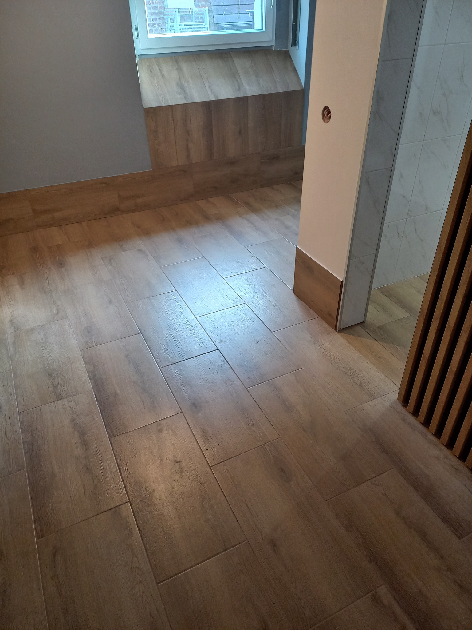 Ułożona pod kątem podłoga z płytek imitujących drewno, z widocznym fragmentem ściany z oknem i wykończeniem w podobnym stylu, oraz fragmentem łazienki z szarymi płytkami i drewnianą dekoracją.