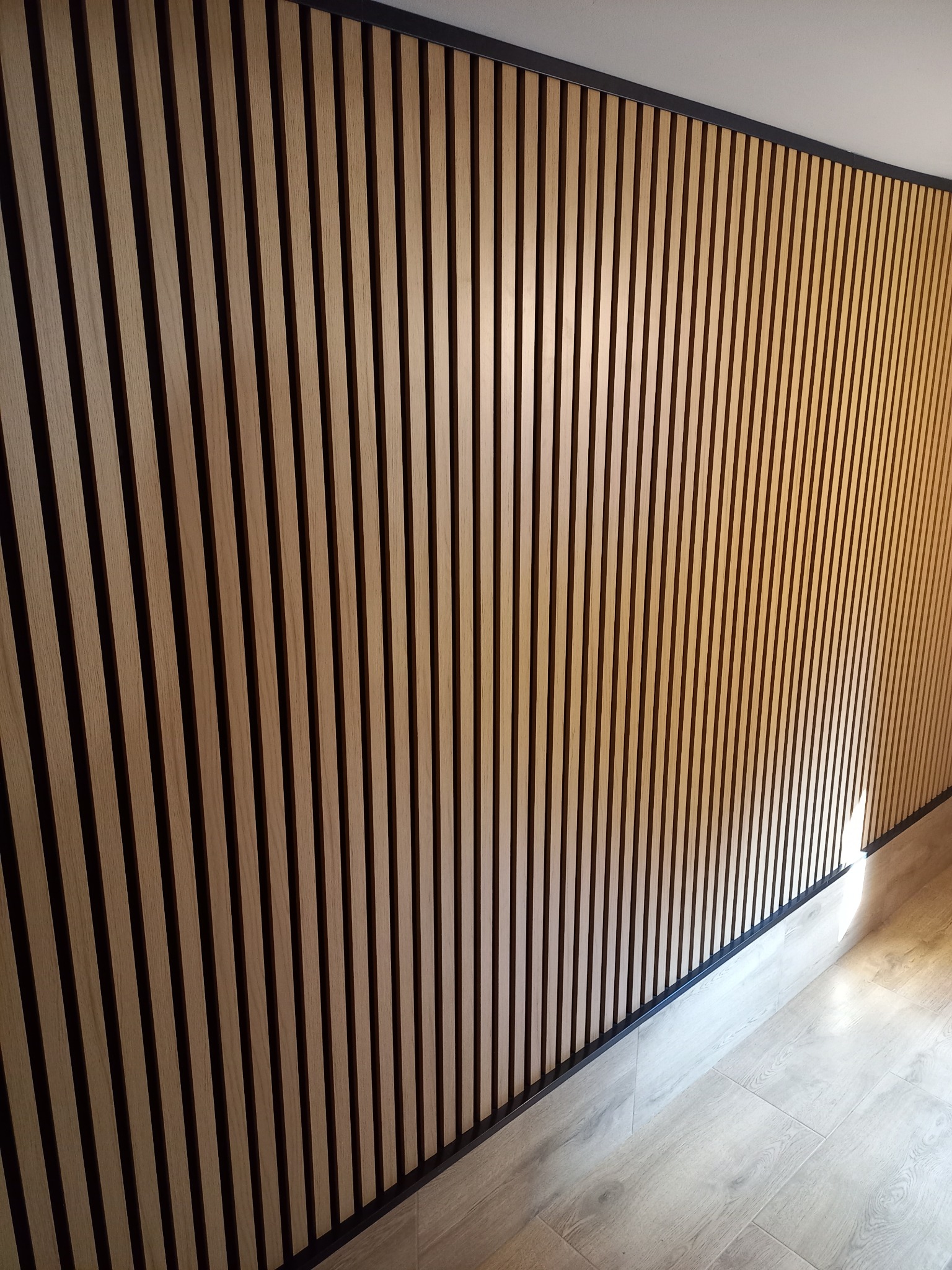 Ściana wykończona panelami lamelowymi w odcieniach naturalnego drewna, z czarnymi akcentami, na podłodze jasne panele imitujące drewno.