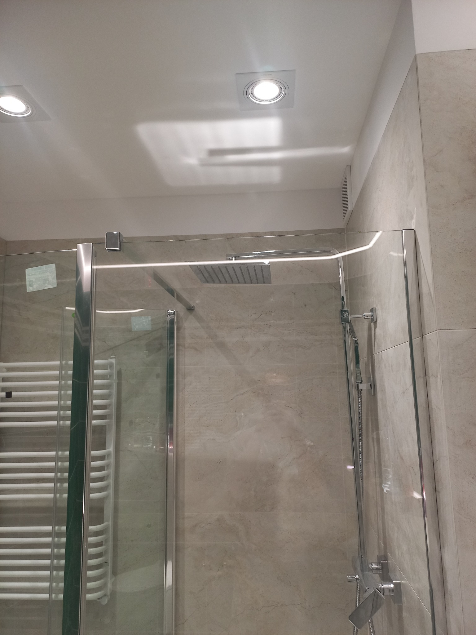 Szklana kabina prysznicowa z deszczownicą i chromowanymi elementami, zamontowana w łazience wykończonej płytkami imitującymi marmur, widoczne oświetlenie sufitowe i drabinkowy grzejnik.