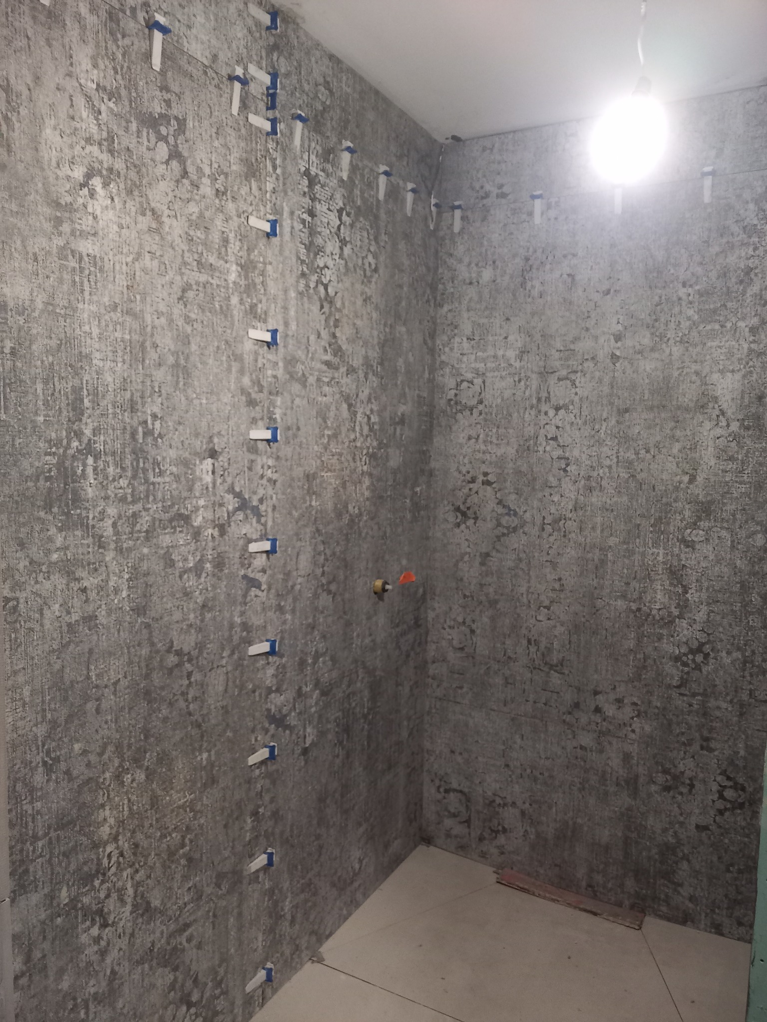 Ściany łazienki w trakcie układania płytek o wzorze imitującym beton, z użyciem systemu poziomowania płytek z niebieskimi klipsami, widoczny fragment instalacji wodnej.