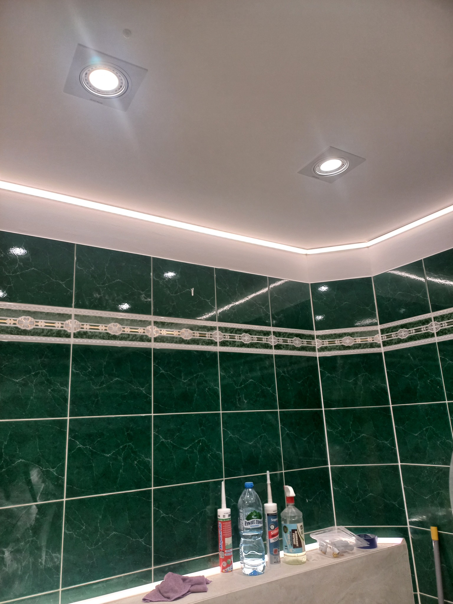 Wykończona łazienka z zielonymi marmurkowymi płytkami, ozdobnym pasem płytek, oświetleniem LED podsufitowym i dwoma kwadratowymi lampami sufitowymi. Na blacie widoczne materiały budowlane i butelka...