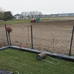 D P KRÓL SPÓŁKA Z OGRANICZONĄ ODPOWIEDZIALNOŚCIĄ - Przygotowanie terenu pod trawnik z rolki: wyrównana ziemia, traktor z siewnikiem w tle, wał do trawy na pierwszym planie, obrzeża rabat z kamykami.