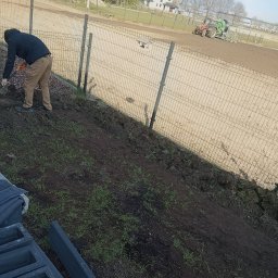 D P KRÓL SPÓŁKA Z OGRANICZONĄ ODPOWIEDZIALNOŚCIĄ - Pracownik w ciemnej kurtce i beżowych spodniach, używając szpadla, przygotowuje ziemię pod nasadzenia wzdłuż metalowego ogrodzenia. W tle traktor na polu.
