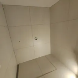 Wykończona kabina prysznicowa z jasnymi, kwadratowymi płytkami, odpływ liniowy w podłodze i wystająca żarówka bez klosza z sufitu.