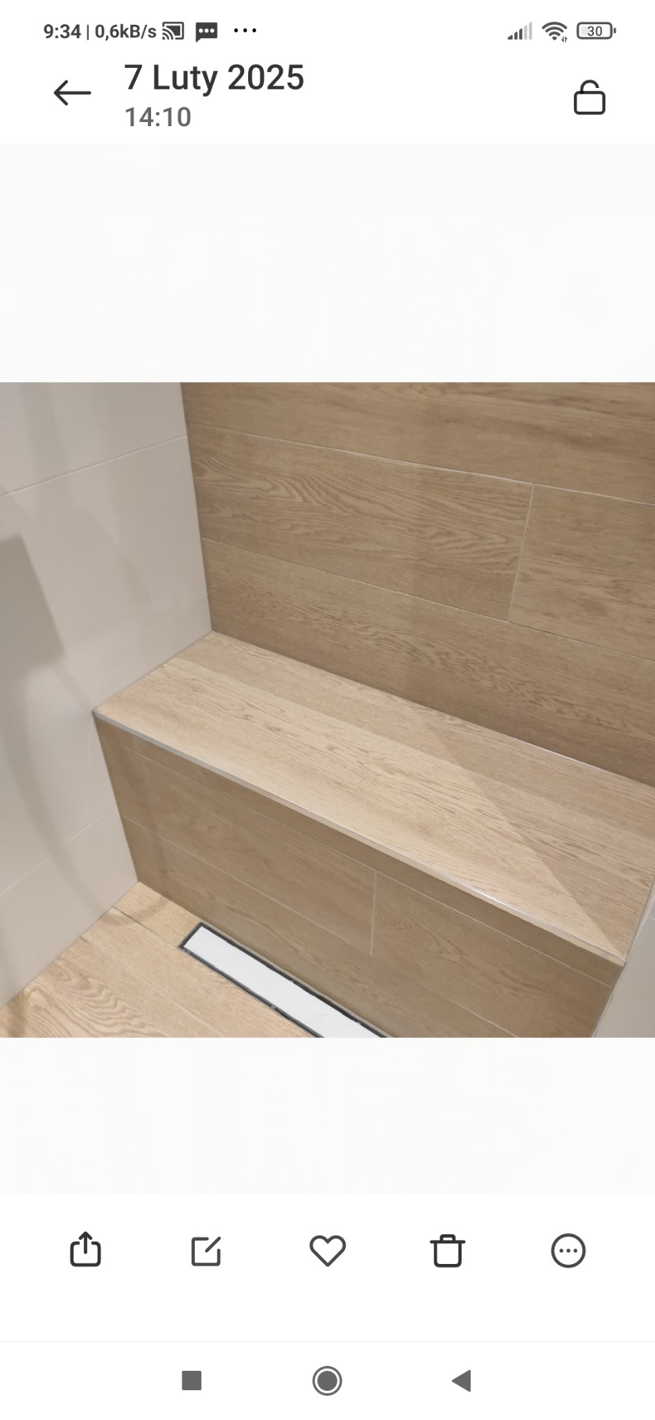 Wykończona łazienka z siedziskiem wyłożonym płytkami imitującymi drewno, odpływ liniowy w podłodze, jasne płytki na ścianie.