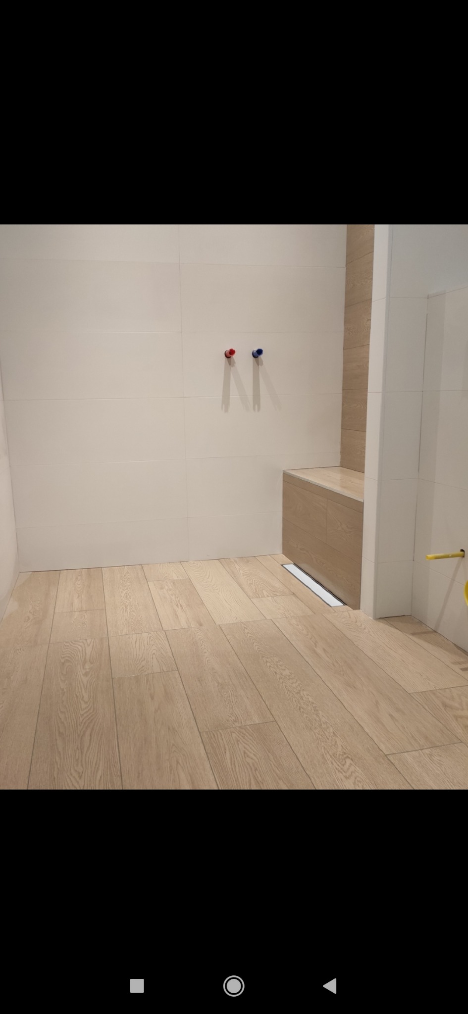 Jasna łazienka z drewnopodobną podłogą i białymi płytkami ściennymi, widoczne przyłącza wodne z czerwoną i niebieską końcówką oraz odpływ liniowy.