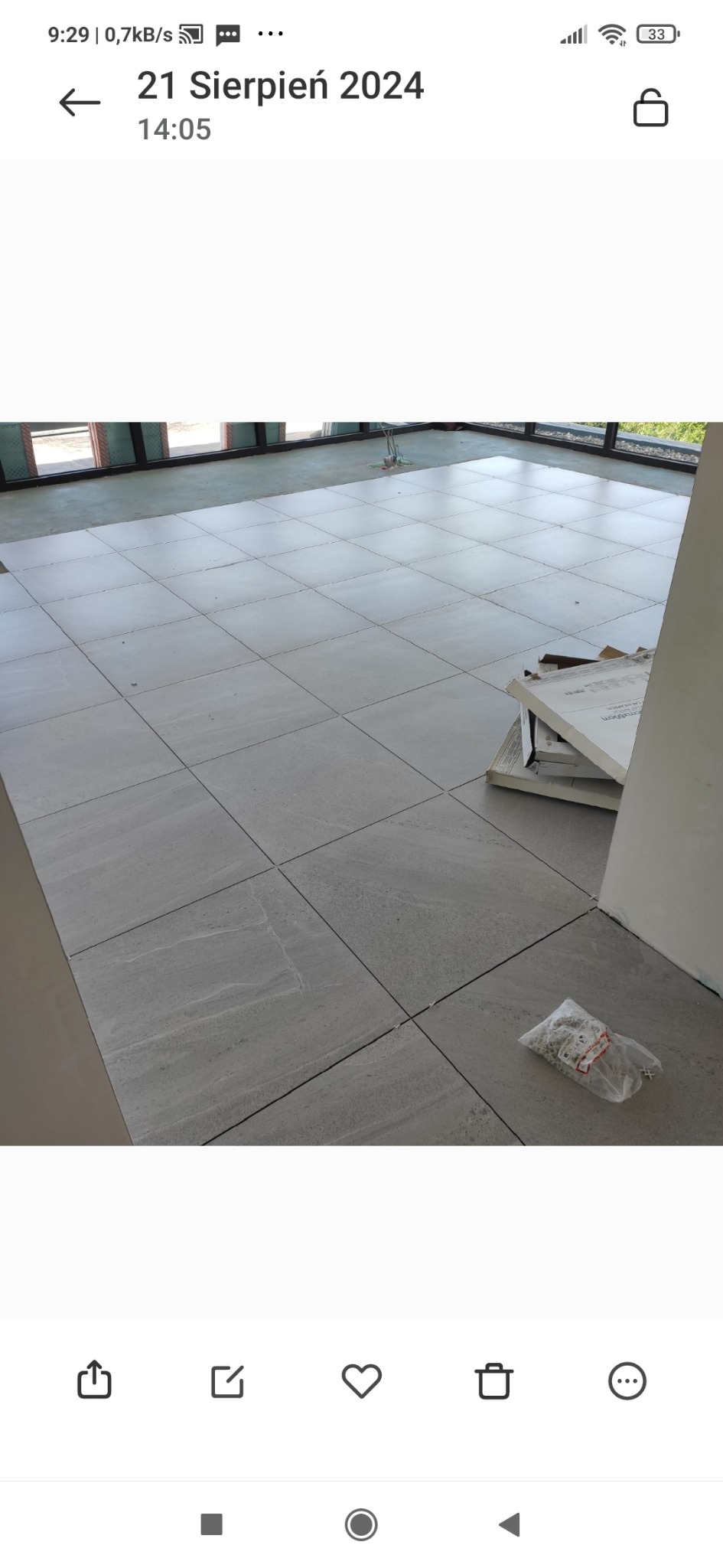 Ułożona podłoga z szarych, kwadratowych płytek w dużym pomieszczeniu z oknami, widoczny worek z krzyżykami dystansowymi i kartonowe opakowania po płytkach.