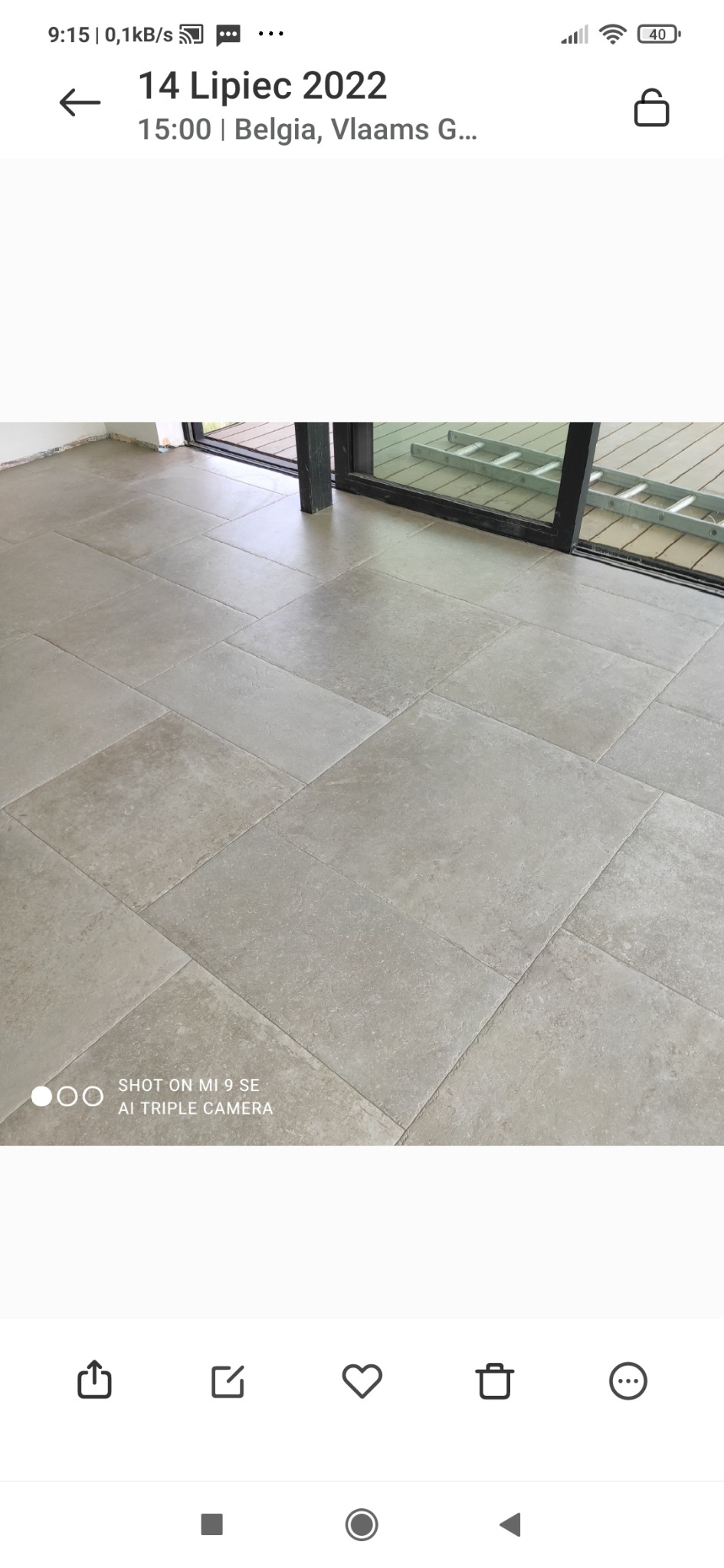 Wyłożona jasnoszarą, matową terakotą podłoga w pomieszczeniu z dużym przesuwnym oknem balkonowym, za którym widoczny jest drewniany taras i aluminiowa drabina oparta o ścianę.