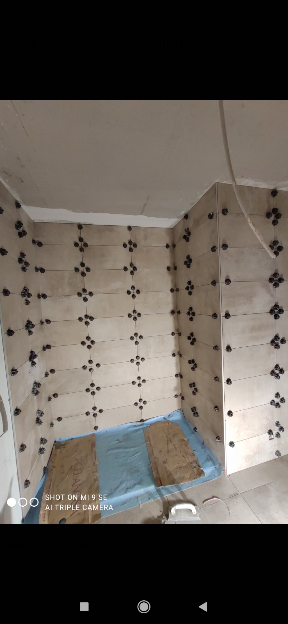 Ściany łazienki w trakcie układania beżowych płytek, widoczne krzyżyki dystansowe, zabezpieczona folią podłoga i narzędzia pracy.