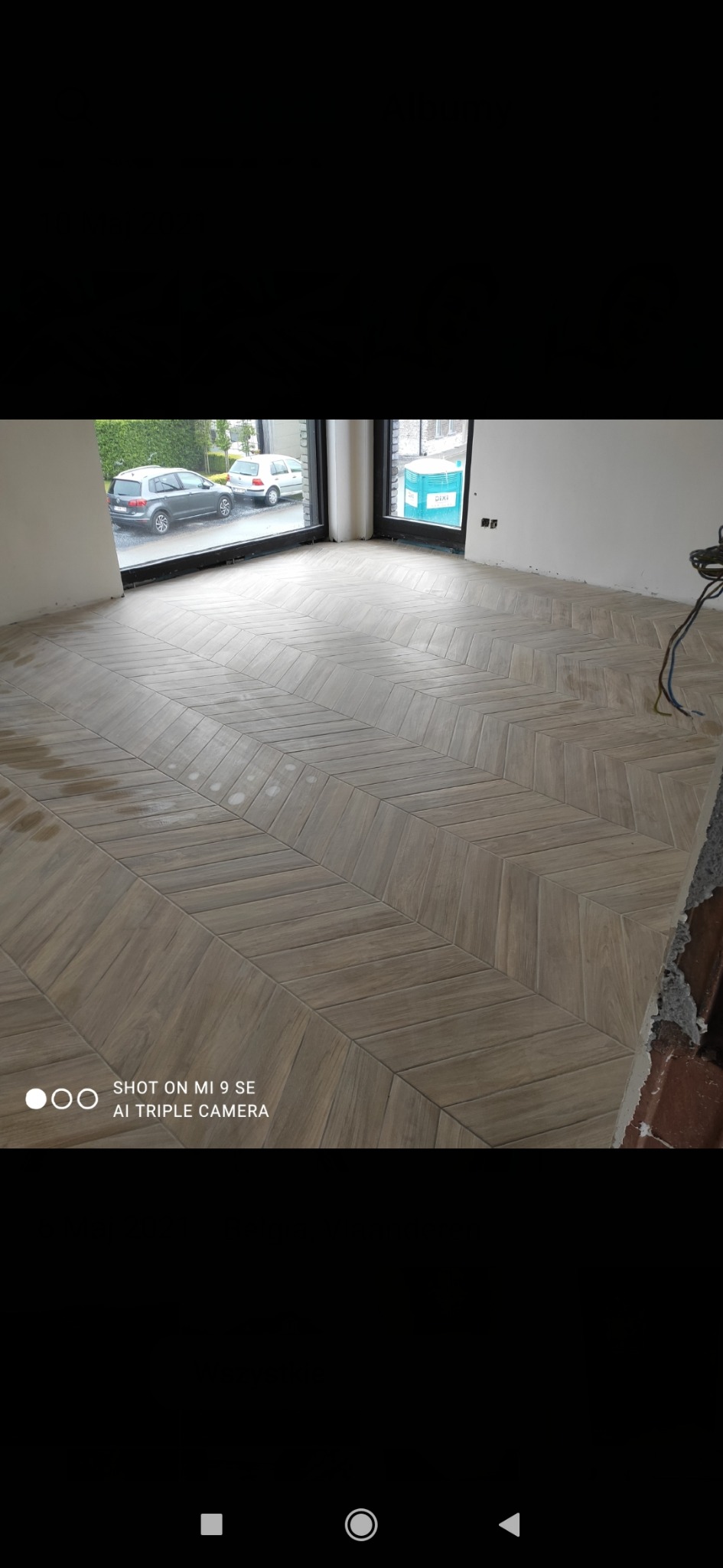 Ułożona pod kątem jasnobrązowa podłoga z płytek imitujących drewno, widok z góry, fragment okna z widokiem na samochody i zieleń, niedokończone ściany.