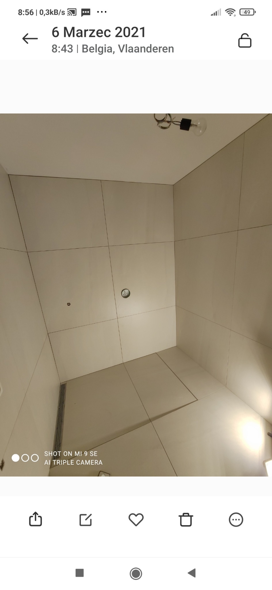 Wykończona kabina prysznicowa z jasnymi, kwadratowymi płytkami, odpływ liniowy w podłodze i wystająca żarówka bez klosza z sufitu.