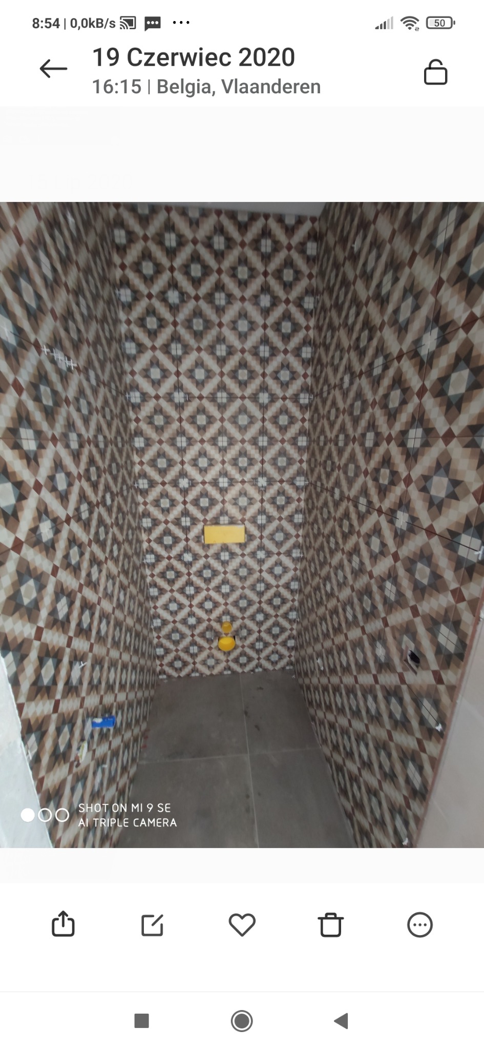 Wnętrze małej łazienki w trakcie wykańczania, ściany wyłożone płytkami w geometryczny wzór, widoczne krzyżyki dystansowe, żółty zawór i niebieski element instalacji na ścianie.