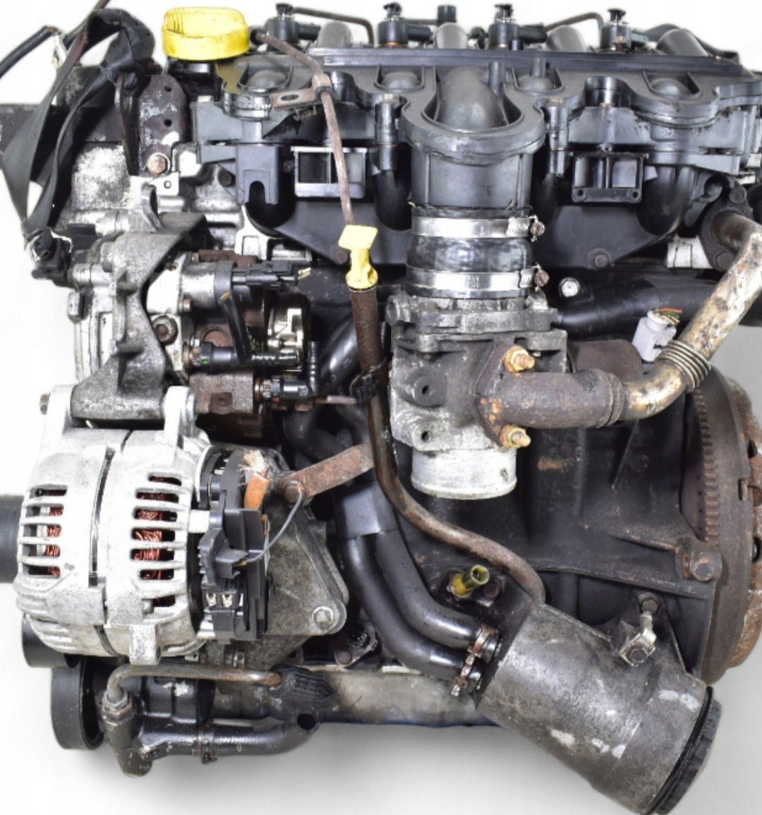Używany, wymontowany silnik samochodowy z widocznymi elementami osprzętu, w tym alternator i przewody, sfotografowany na białym tle.