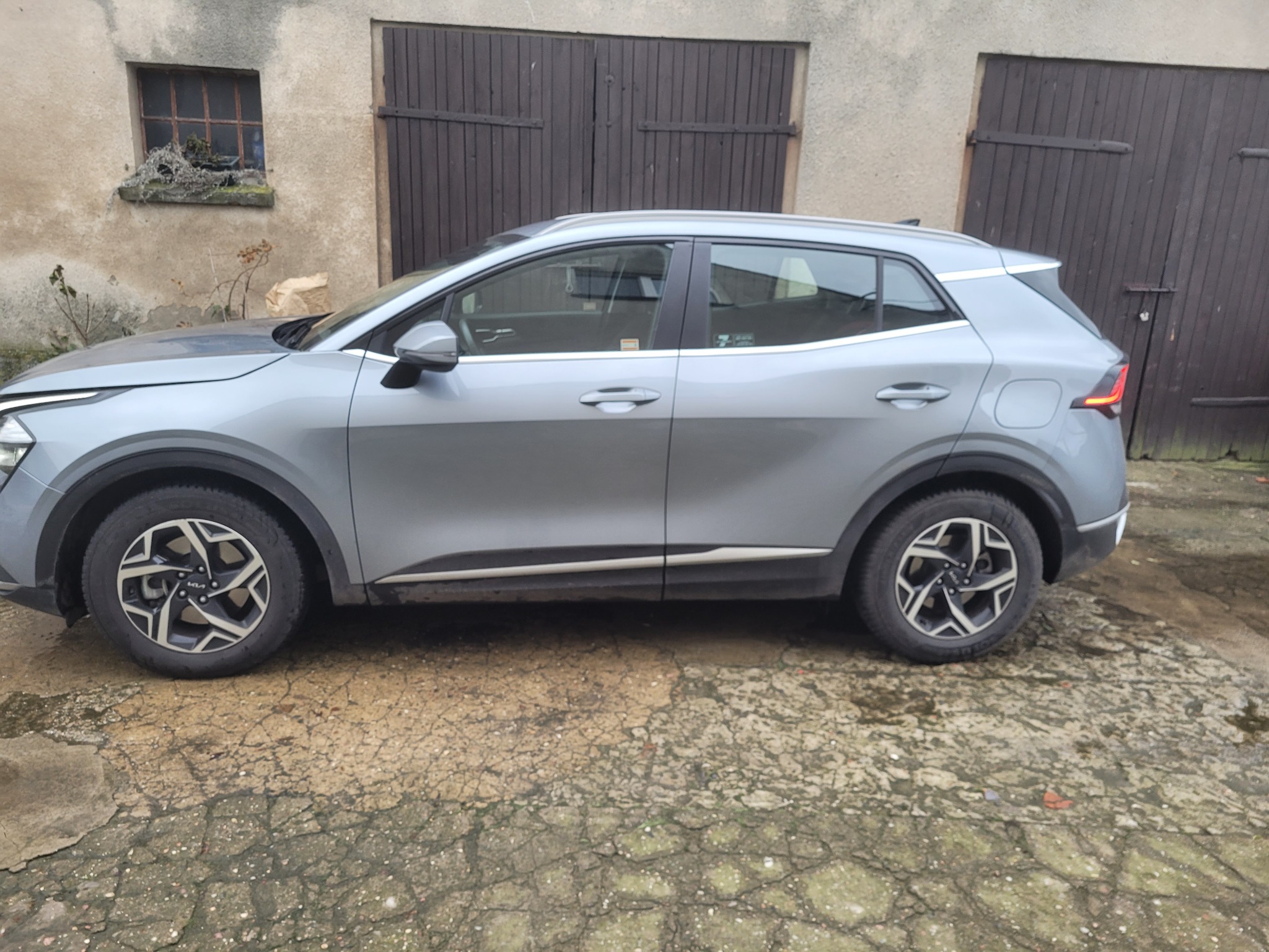 Szary samochód Kia Sportage zaparkowany przed budynkiem z ciemnymi, drewnianymi wrotami garażowymi i małym oknem z roślinami.