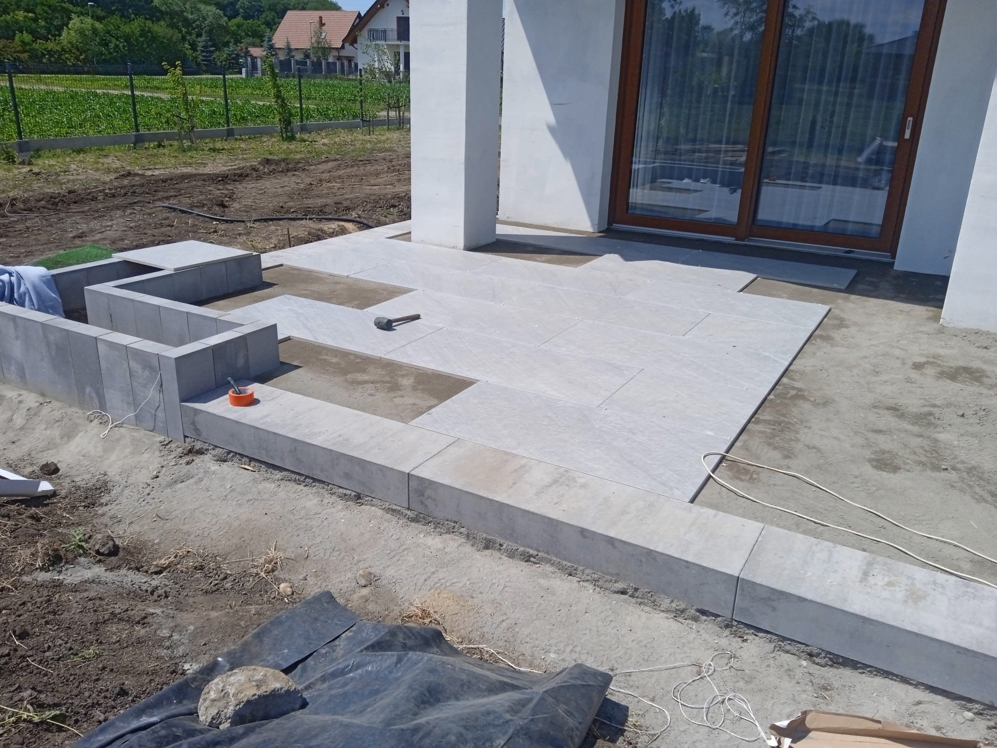 Montaż tarasu z jasnych płyt kamiennych, widoczne niedokończone fragmenty i gumowy młotek, obrzeża z betonowych bloczków, w tle dom jednorodzinny.
