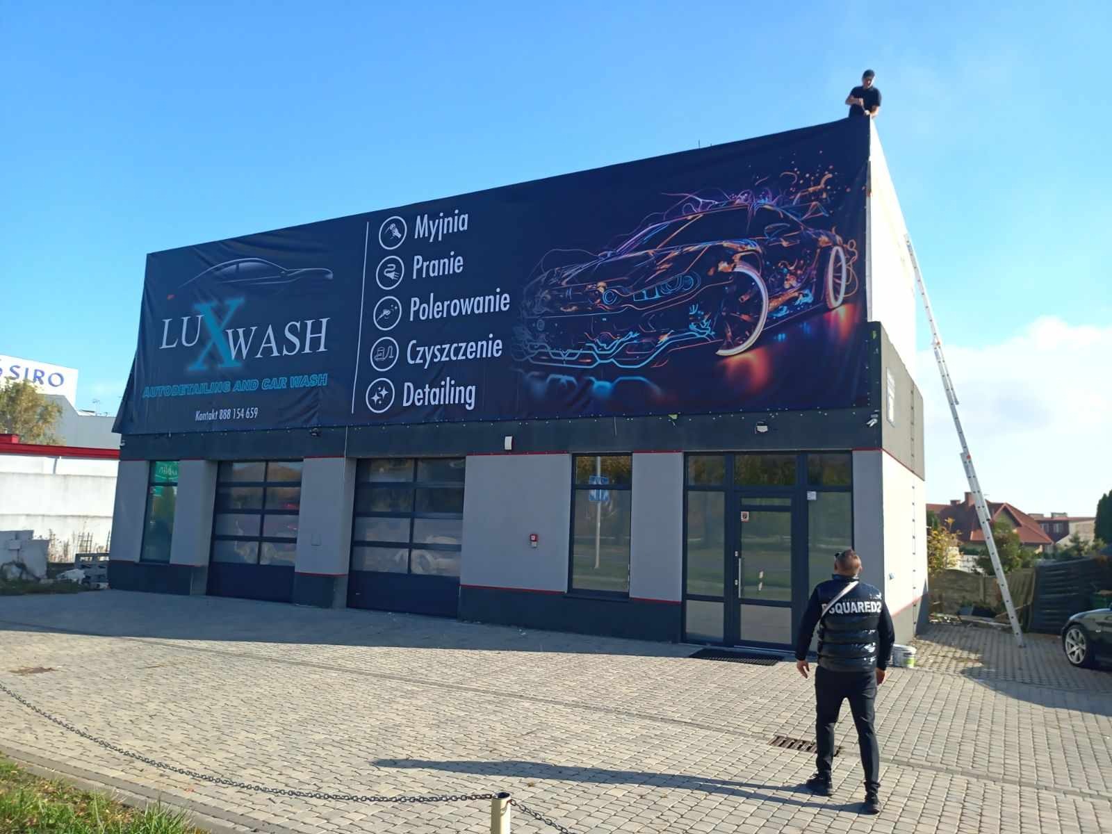 Montaż baneru reklamowego myjni samochodowej LUX WASH z ofertą myjni, prania, polerowania, czyszczenia i detailingu aut. Pracownik na drabinie i drugi na chodniku obserwują proces.