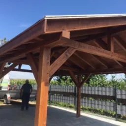 Ciemnobrązowa, drewniana konstrukcja altany z widocznym dachem i słupami, na tle błękitnego nieba i zieleni. Widoczny fragment ogrodzenia i osoby w tle.