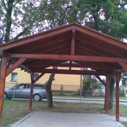 Zakład Stolarski Joachim Michna - Ciemnobrązowa, drewniana altana z widocznym zadaszeniem i słupami, na trawiastym terenie, z drzewem rosnącym wewnątrz konstrukcji. W tle żółty budynek i szary samochód.