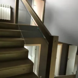 Drewniane schody z nowoczesną szklaną balustradą i drewnianą poręczą. Widoczne trzy stopnie i fragment ściany w szarym odcieniu. Minimalistyczny design.