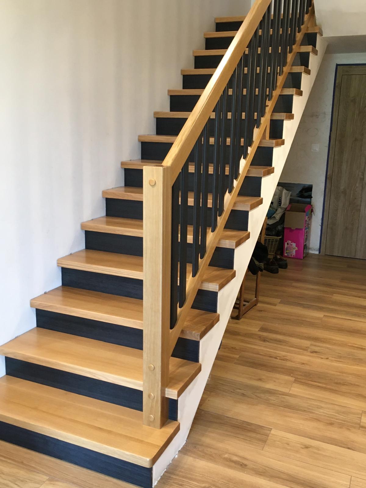 Nowoczesne schody z jasnego drewna z czarnymi podstopnicami i balustradą z prostych, czarnych elementów. Widok z dołu na bieg schodów.