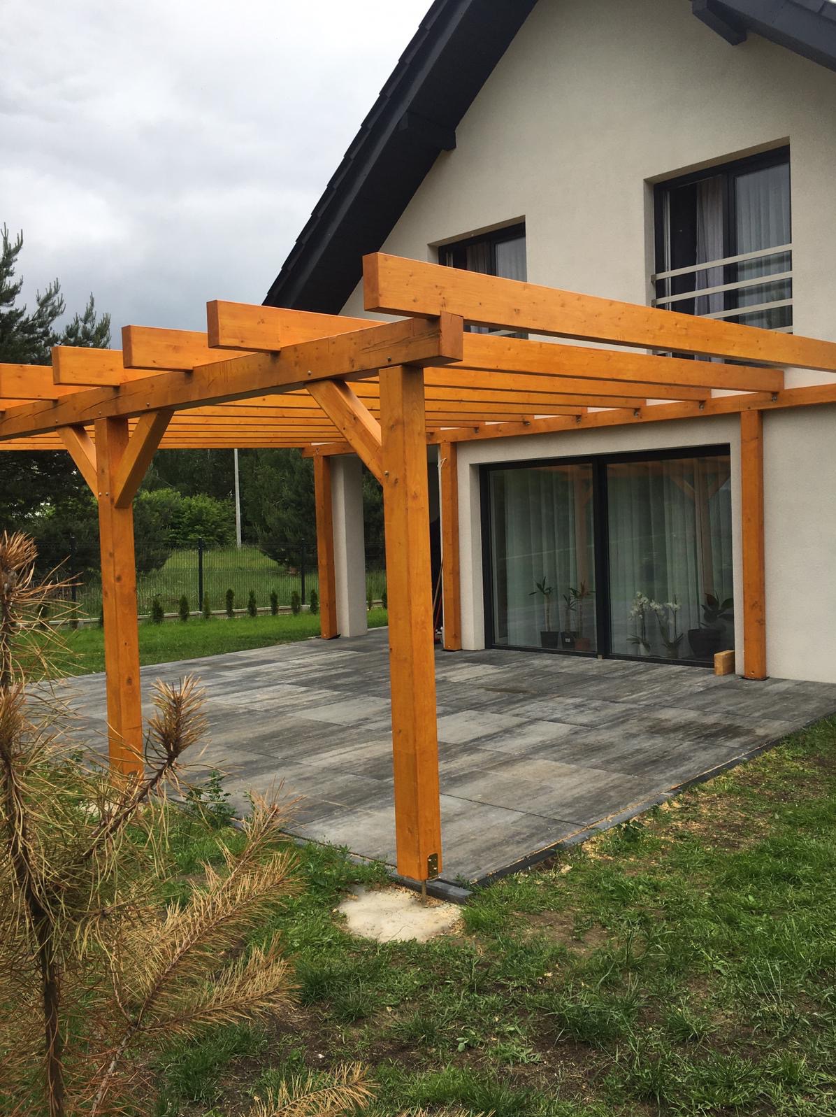 Nowoczesna, jasna pergola z belek na tarasie z szarej kostki, przylegająca do domu z dużymi oknami przesuwnymi.