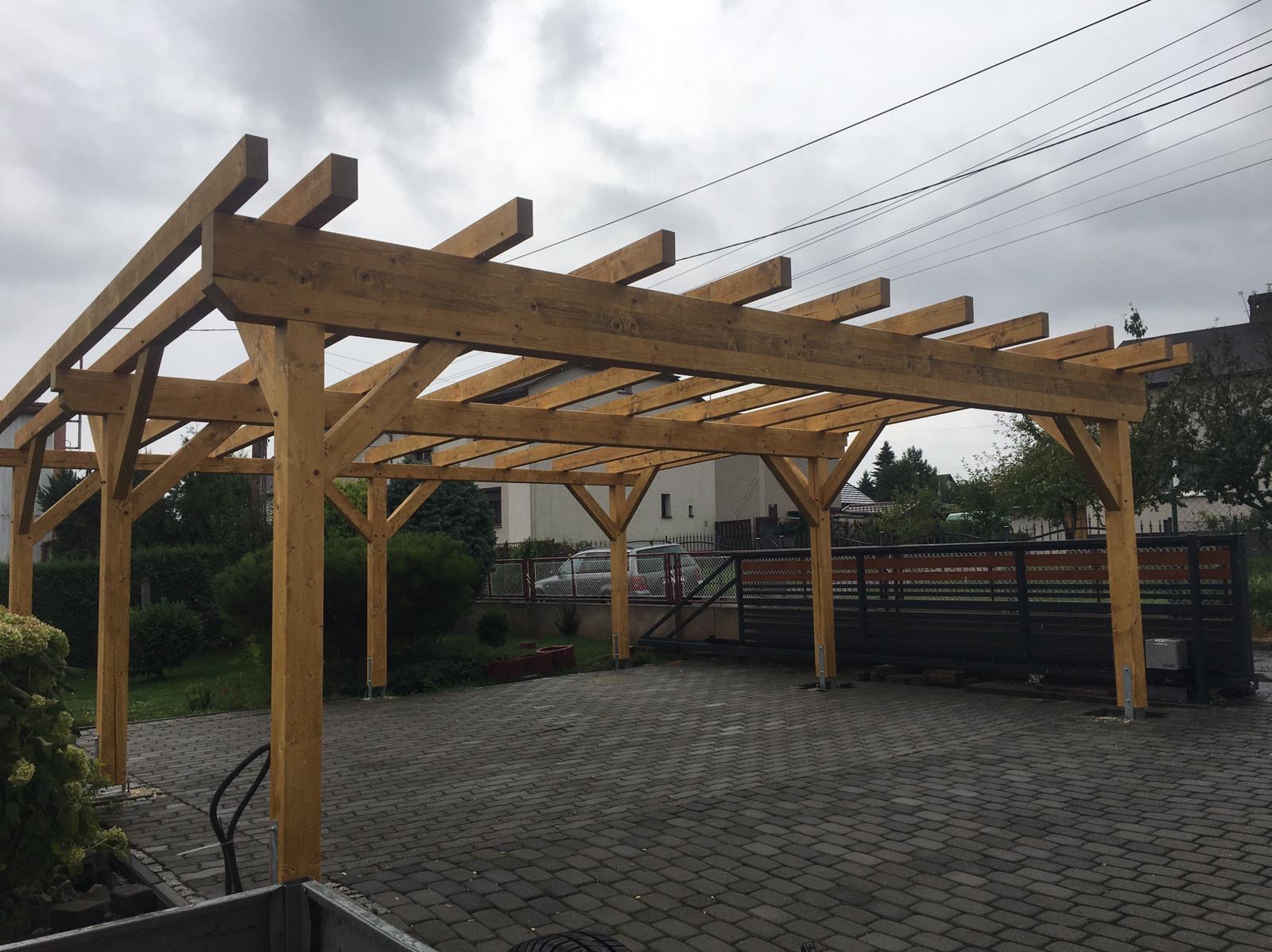 Surowa drewniana pergola w trakcie budowy, widok z dołu na konstrukcję wspartą na słupach. Tło: dom, brama i kostka brukowa.