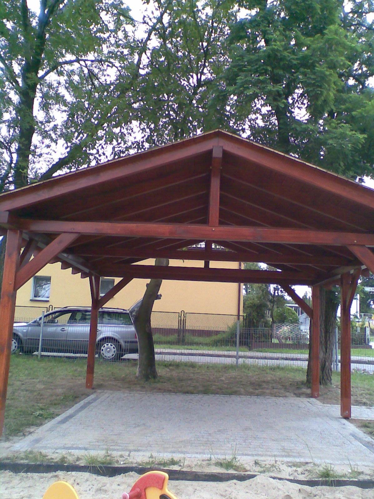 Ciemnobrązowa, drewniana altana z widocznym zadaszeniem i słupami, na trawiastym terenie, z drzewem rosnącym wewnątrz konstrukcji. W tle żółty budynek i szary samochód.