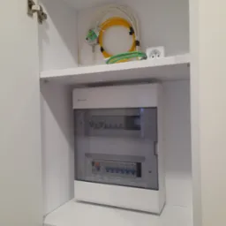 Biała szafka z rozdzielnicą elektryczną Elektro-Plast, widoczne kable światłowodowe i gniazdko elektryczne na górnej półce.