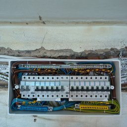 FHU Norbert Winczewski - Otwarta skrzynka rozdzielcza z bezpiecznikami Legrand, widoczne kable w różnych kolorach, zamontowana na nierównej ścianie z widocznymi śladami po instalacji.