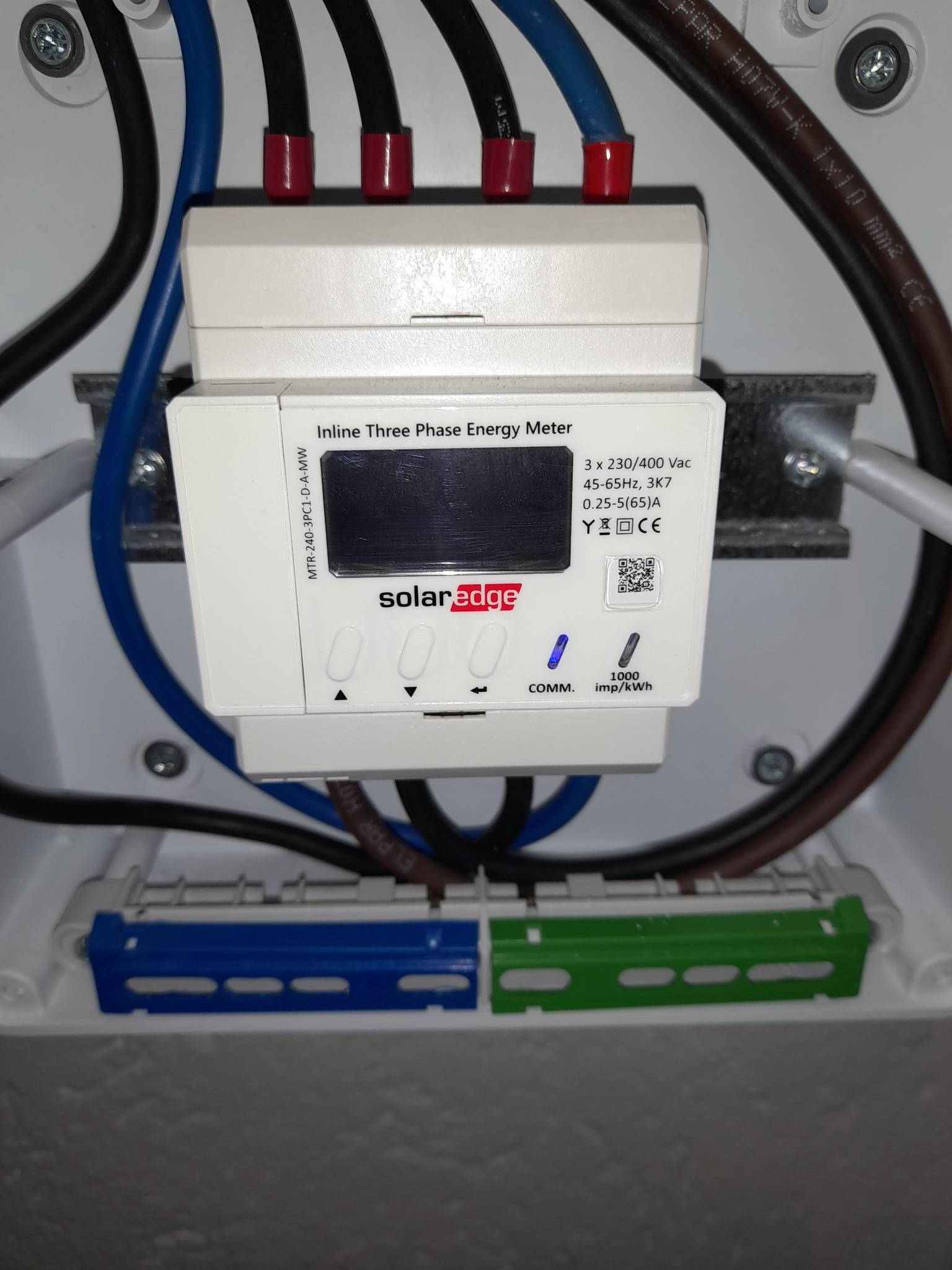 Trójfazowy licznik energii SolarEdge zamontowany w skrzynce elektrycznej z widocznymi kablami w kolorach niebieskim, czarnym i brązowym, podłączony do listew zaciskowych.