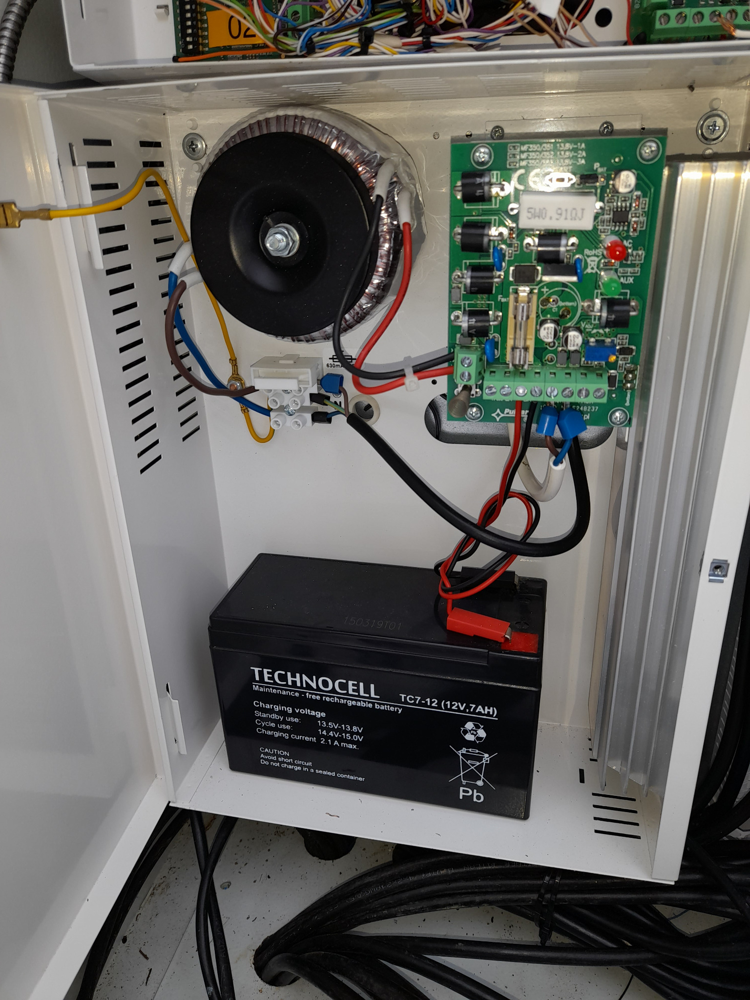 Wnętrze białej szafki sterowniczej z akumulatorem Technocell 12V, transformatorem toroidalnym i płytką drukowaną z elementami elektronicznymi, widoczne okablowanie i złącza.